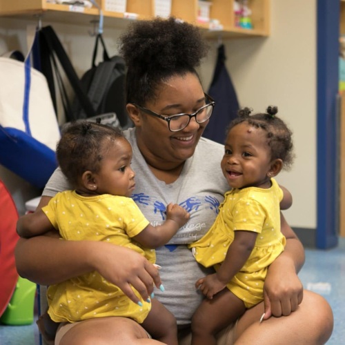 A woman holds two smiling babies wearing yellow jumpsuits in a classroom.